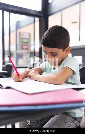 A scuola, in classe, giovane ragazzo birazziale che si concentra sulla scrittura su un taccuino Foto Stock