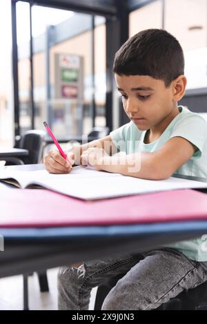 A scuola, in classe, giovane ragazzo birazziale che si concentra sulla scrittura su un taccuino Foto Stock