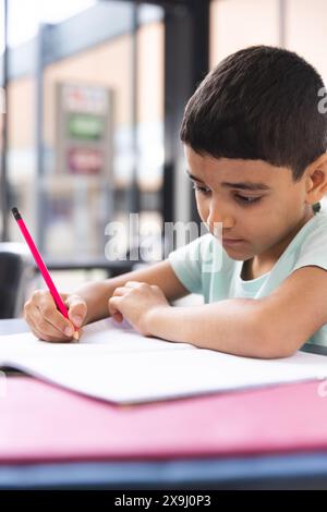 A scuola, giovane ragazzo birazziale che si concentra sulla scrittura in un taccuino in classe Foto Stock