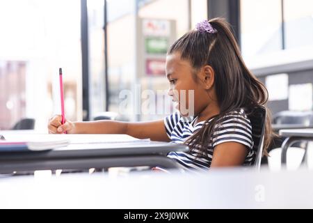 A scuola, giovane ragazza birazziale con pelle marrone chiaro sta scrivendo in classe Foto Stock