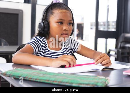 A scuola, giovane ragazza birazziale che indossa le cuffie e si concentra sulla lettura in classe Foto Stock