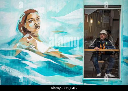 Una persona all'interno si siede in una caffetteria accanto a una nuova opera d'arte graffiti presso l'Upfest presenta un evento culturale, una Street art e una celebrazione dei graffiti nella zona di Bedminster di Bristol. Si svolge per diciassette giorni, con celebri artisti di strada provenienti da tutto il mondo ed è il più grande festival di Street art e graffiti d'Europa. Data foto: Sabato 1 giugno 2024. Foto Stock