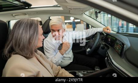 Un uomo anziano tiene le chiavi mentre è seduto in una macchina nuova. Una coppia sposata dai capelli grigi è felice di comprare un'auto. Foto Stock