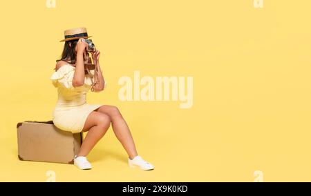 un abito e un cappello ingiallo da donna, che porta con sé una valigia e scatta foto con una macchina fotografica vintage su sfondo giallo. Gente felice che va in vacanza, libera Foto Stock