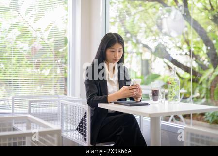 Donna d'affari professionale che lavora sul suo smartphone in un bar luminoso e moderno con caffè e vegetazione Foto Stock