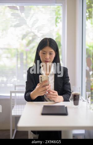 Donna d'affari asiatica professionista che lavora sul suo smartphone in un bar luminoso e moderno con caffè e vegetazione Foto Stock