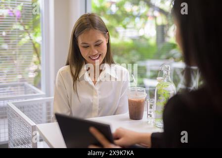 Due donne asiatiche hanno una piacevole conversazione con un caffè in un moderno ambiente da caffe' con vegetazione esterna. Foto Stock