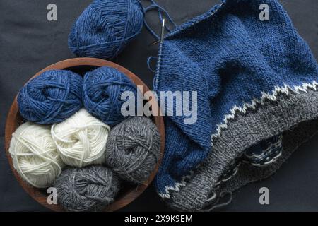 Fotografia piatta di stockinette di lana bianca grigia blu con cuciture a maglia su un ago di metallo con un cestino con palle di filato e forbici vintage, lavorate a maglia Foto Stock