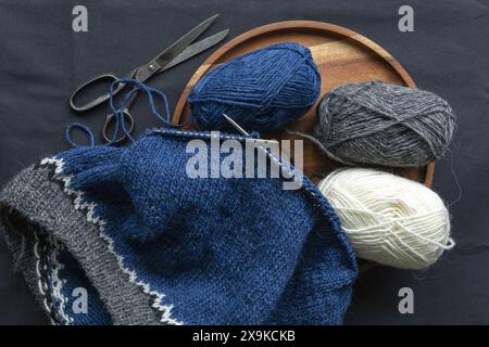 Fotografia piatta di un maglione in maglia di lana bianca grigia blu sugli aghi in metallo con palle in filato e forbici vintage, maglieria con cuciture stockinette Foto Stock