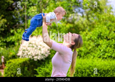 Una scena gioiosa di una madre che solleva il suo bambino ridendo in alto nell'aria mentre gioca in giardino. Foto Stock