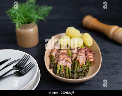 Asparagi verdi cotti avvolti nella pancetta e serviti con patate bollite su splendidi piatti beige su sfondo scuro Foto Stock