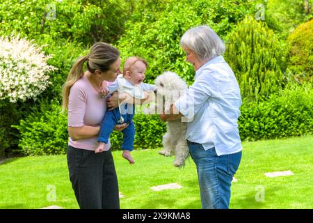 Un momento di gioia catturato mentre tre generazioni si riuniscono in un giardino lussureggiante. Una giovane madre tiene il suo bambino, che sta raggiungendo un morbido cane bianco Foto Stock