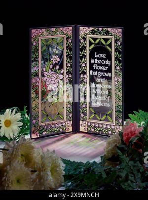 Tessera pieghevole in vetro colorato con motivi floreali e citazione d'amore ispiratrice, circondata da fiori morbidi su sfondo scuro, romantica decorazione a. Foto Stock