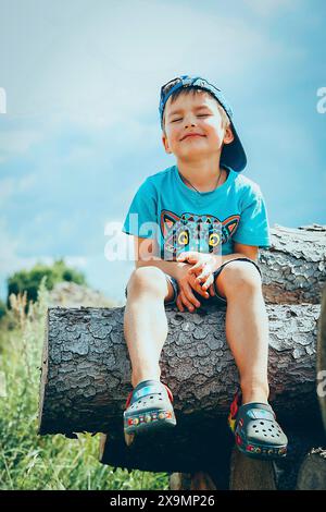 Ragazzo seduto su un tronco all'aperto in una giornata di sole, con una camicia e un berretto blu e scarpe colorate, Bielorussia, Minsk Foto Stock