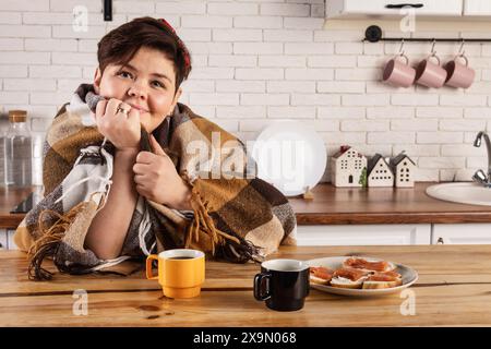 Una donna in sovrappeso siede al suo tavolo da cucina, avvolto in un'accogliente coperta, gustando una tazza di caffè e un piatto di deliziosi panini. Sembra squallida Foto Stock