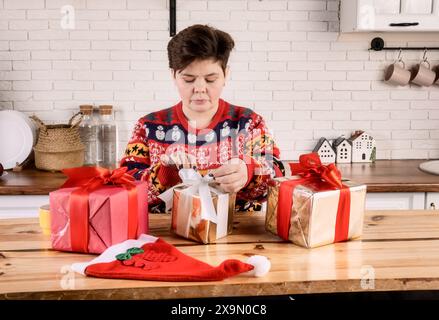 Donna che avvolge i regali di Natale in carta rossa e oro con nastro abrasivo. Due regali confezionati, uno parzialmente avvolto, cappello di Babbo Natale rosso sul tavolo. Donna in festa Foto Stock