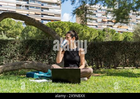 Una donna si diverte a lavorare in remoto in un parco tranquillo, utilizzando un computer portatile e gustando un caffè in una giornata di sole Foto Stock