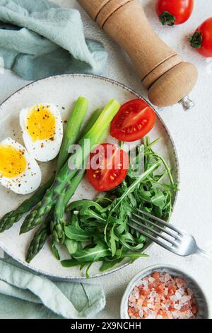 Una sana insalata primaverile si trova su un tavolo bianco, con uova bollite a fette, asparagi verdi vivaci e pomodori ciliegini maturi insieme a rucola fresca Foto Stock