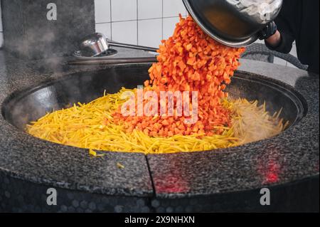 Lo chef maschile aggiunge carote a un calderone per cucinare il tradizionale pilaf uzbeko in cucina in un ristorante in Uzbekistan Foto Stock