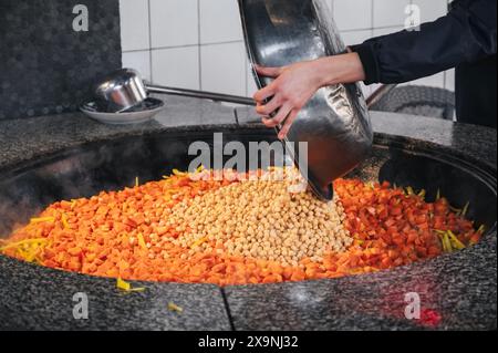 Lo chef maschile aggiunge ceci al calderone con carote per cucinare il tradizionale pilaf uzbeko orientale in cucina in un ristorante dell'Uzbekistan Foto Stock
