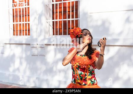 Una donna vestita con un vivace abito arancione con motivi floreali e un capo coordinato applica il trucco con uno specchio compatto. E' di fronte a un bianco Foto Stock