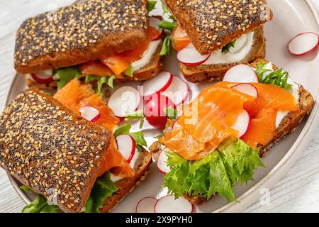 panini di salmone affumicati su panini di segale con ravanello rosso fresco, lattuga e panna freschi sul piatto su un tavolo di legno bianco, primo piano Foto Stock