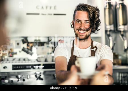 Bel barista che lavora in caffetteria, preparando il caffè per il cliente. Studente universitario che lavora part-time in un bar. Foto Stock