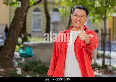 Tipo. Felice uomo asiatico di mezza età alza il pollice in su concorda con qualcosa che dà risposta positiva consiglia pubblicità feedback di buone idee, celebrando la vittoria di successo all'aperto. Ragazzo sulla strada della città. Foto Stock