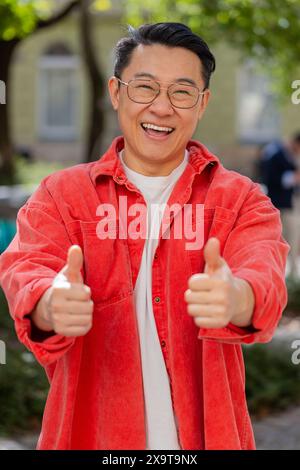 Tipo. Felice uomo asiatico di mezza età alza il pollice in su concorda con qualcosa che dà risposta positiva consiglia pubblicità feedback di buone idee, celebrando la vittoria di successo all'aperto. Ragazzo sulla strada della città. Foto Stock