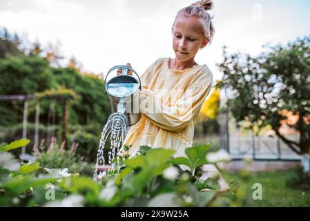 Ragazza che annaffia fragole in un letto rialzato e tiene una lattina di metallo. Prendersi cura dell'orto e piantare frutta, verdura e fiori primaverili. Foto Stock