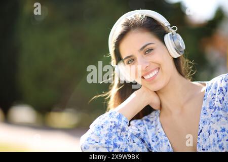 Una donna felice che indossa le cuffie guarda la fotocamera seduta in un parco Foto Stock