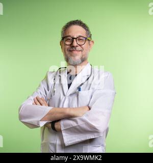 Medico sorridente con barba e occhiali, camice da laboratorio e stetoscopio, in piedi con le braccia incrociate, sfondo verde chiaro Foto Stock