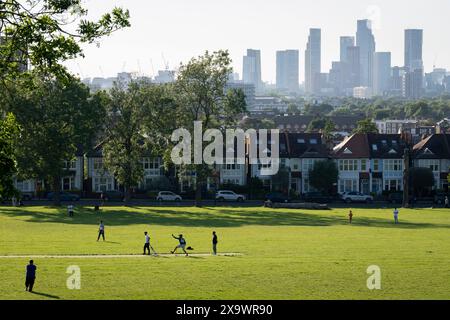 I giocatori dilettanti di cricket godono della luce del sole della domenica sera sullo sfondo di uno skyline a sud di Londra di grattacieli a Nine Elms a Battersea, visto da Ruskin Park, uno spazio verde pubblico a Lambeth, il 2 giugno 2024, a Londra, Inghilterra. Foto Stock