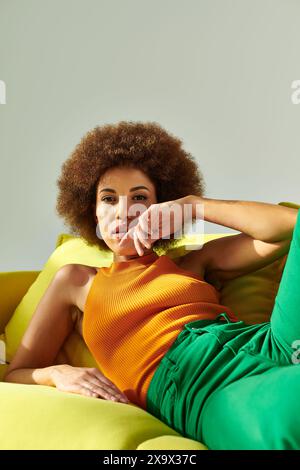 Una donna riccia afro-americana siede su un divano giallo brillante, che mostra relax. Foto Stock