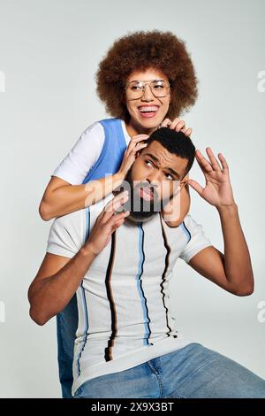 Un elegante afroamericano si siede giocosamente sulla testa di una donna su uno sfondo grigio, mostrando amicizia e fiducia. Foto Stock