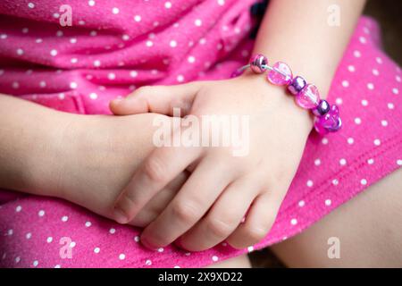 Mani da bambina con bracciale in primo piano, vestito con un abito a pois rosa, che crea un bracciale personalizzato con perline colorate, catturando fascino e delicatezza Foto Stock