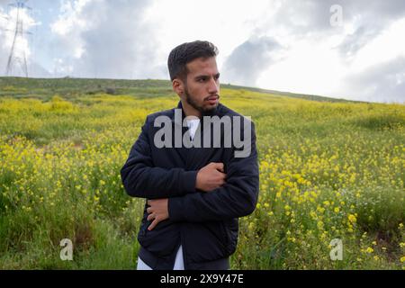 Un uomo con una giacca nera e un disdasha si trova in un campo vibrante di fiori gialli e verdi colline, braccia incrociate per il calore, sotto un cielo primaverile nuvoloso, Foto Stock