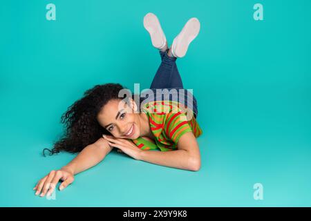 Foto a figura intera di una bella ragazza sdraiata sul pavimento, vestito da sogno, elegante abito a righe isolato su sfondo di colore ciano Foto Stock