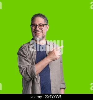 Uomo maturo con occhiali e barba, sorridente e rivolto lateralmente, indossa un abito casual, una camicia blu e una giacca beige, in piedi contro una b verde Foto Stock