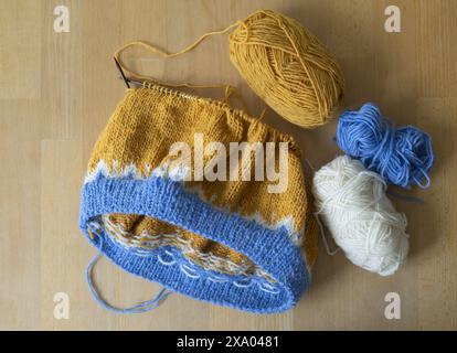 Fotografia piatta di un maglione in lopapeysa lavorato a maglia di lana islandese in corso con palline in filato giallo, blu e bianco e aghi circolari su un WO Foto Stock