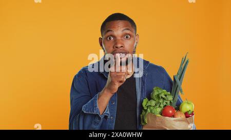 Un ragazzo afroamericano segreto che fa un gesto muta con il dito sulle labbra, mantenendo un segreto sui prodotti biologici coltivati localmente in un sacchetto di carta. Una persona privata sul veganismo. Telecamera A.. Foto Stock