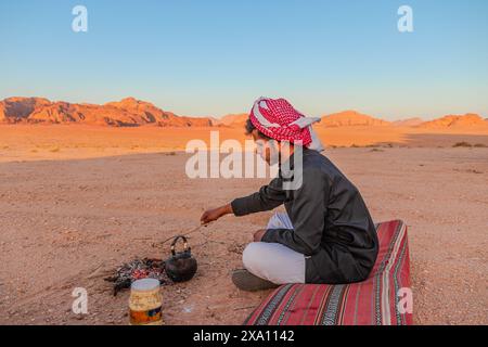 Giordania - 1° maggio 2024: L'uomo beduino tradizionale prepara il tè su un fuoco nella vasta distesa del deserto del Wadi Rum giordano mentre il sole tramonta Foto Stock