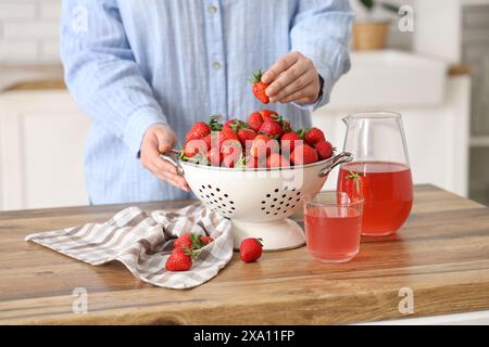 Donna con fragole fresche nel colino e succo di fragole su un tavolo di legno in cucina Foto Stock