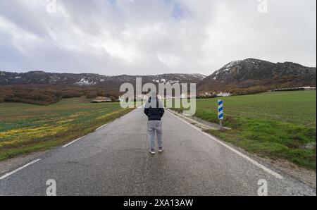 Un uomo cammina lungo una strada umida che conduce a un villaggio di Navarra, in Spagna, mentre guarda le montagne innevate e il cielo coperto. Valle Baztan Foto Stock