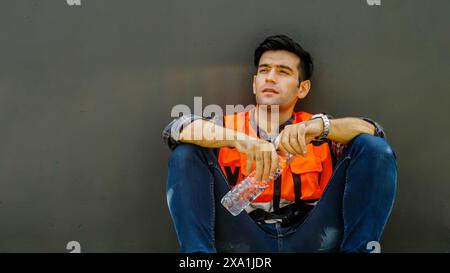 Un operaio edile che regge una bottiglia d'acqua, appoggiato contro un muro grigio Foto Stock