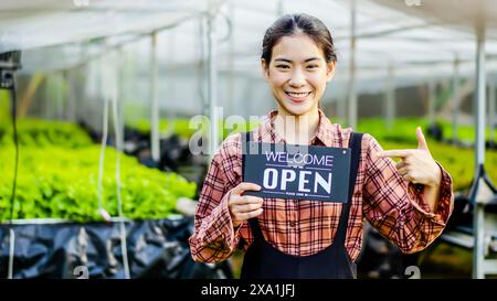 Una donna asiatica sorridente che lavora in una serra, con un cartello di benvenuto Foto Stock