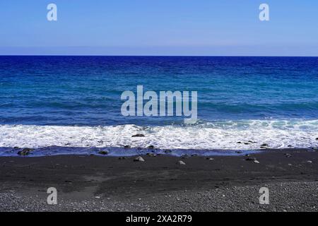 Tenerife, Isole Canarie, Spagna, Europa, Vista di una spiaggia calma con onde che colpiscono delicatamente la sabbia nera sotto un cielo azzurro Foto Stock