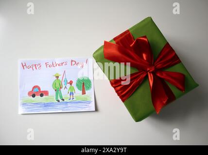 Foto della festa del papà di una cartolina disegnata da un bambino. Un regalo di colore verde con un fiocco rosso. Sfondo bianco. La cartolina mostra un padre e un figlio a pescare. Aspetto africano. Disegno con matite colorate. Foto Stock