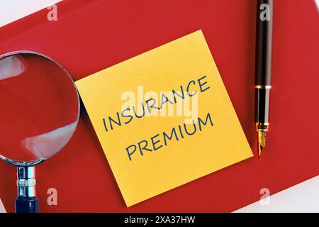 Concetto finanziario. PREMIO ASSICURATIVO su un adesivo giallo su un quaderno rosso in una composizione con una lente d'ingrandimento e una penna stilografica Foto Stock