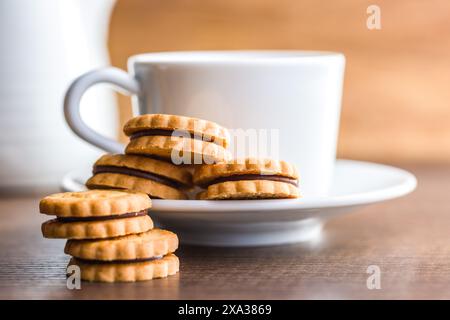 Immagine ravvicinata di una pila di biscotti ripieni di cioccolato poggiati su un tavolo di legno. Una tazza bianca e una caraffa sono visibili sullo sfondo, fuori fuoco Foto Stock